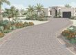 A curved gray paver driveway leads to a Florida home among palms and flowers.