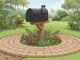 A roadside mailbox surrounded by circular pavers, small flowers, and tropical greenery.
